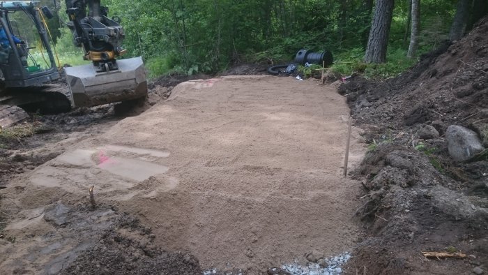 Byggnation av markbädd med grävmaskin, avjämnad sand och rör vid skogsområde.