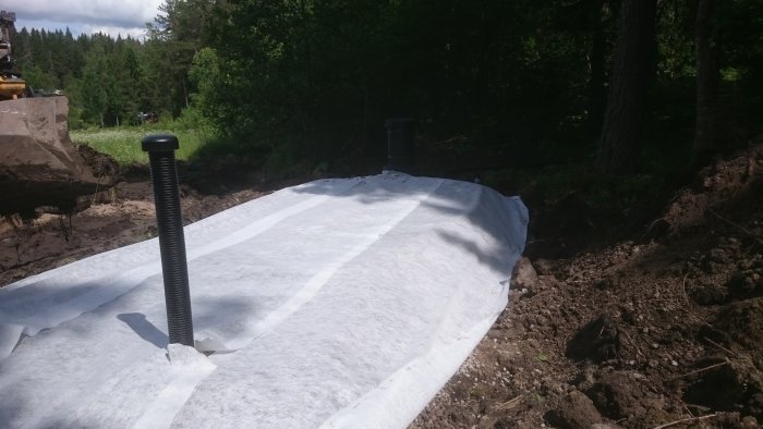 Markbädd under konstruktion med geotextil och ventilerad rörstång framför skogsmark.