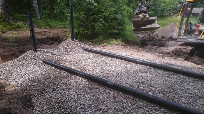 Byggande av markbädd med grus och rör, grävmaskin i bakgrunden på en arbetsplats.
