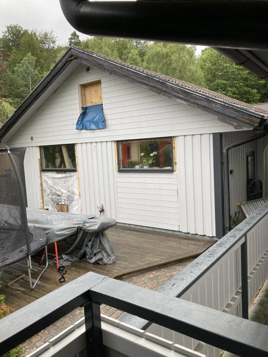 Husfasad under renovering med omonterat fönster täckt av blå presenning, veranda i förgrunden.