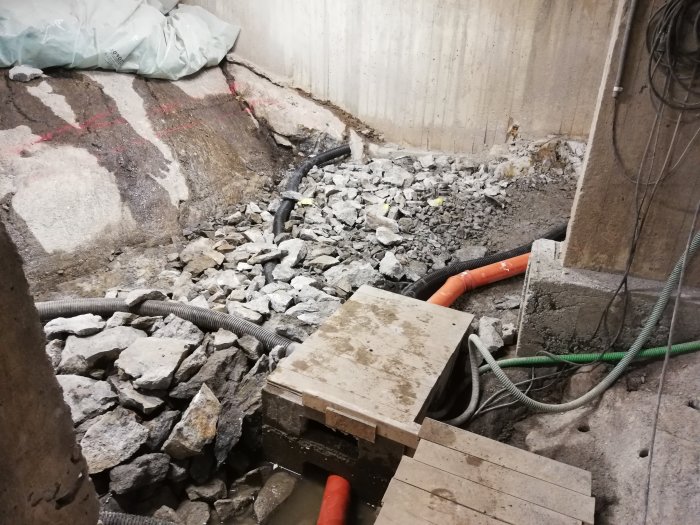 En delvis återfylld byggarbetsplats med provmonterade dräneringsrör, varav ett långt orange, i en grusig och stenig miljö.