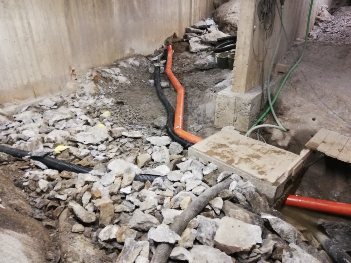 Orange dräneringsrör provmonterade i en utgrävd byggarbetsplats med grus och sten, betongväggar och trästöd.