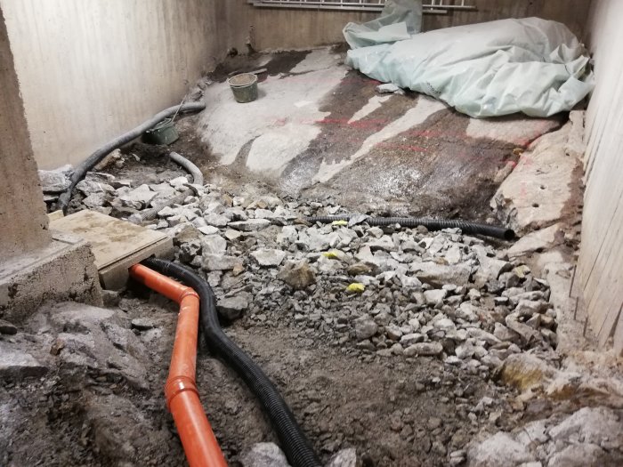 Ett delvis återfyllt källarutrymme med provmonterat orange dräneringsrör och svart slang mot en betongvägg.