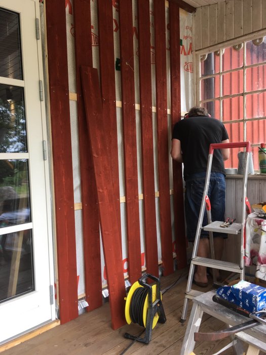 Person monterar rödmålad panel på vägg med vindskydd bakom och byggmaterial i förgrunden.