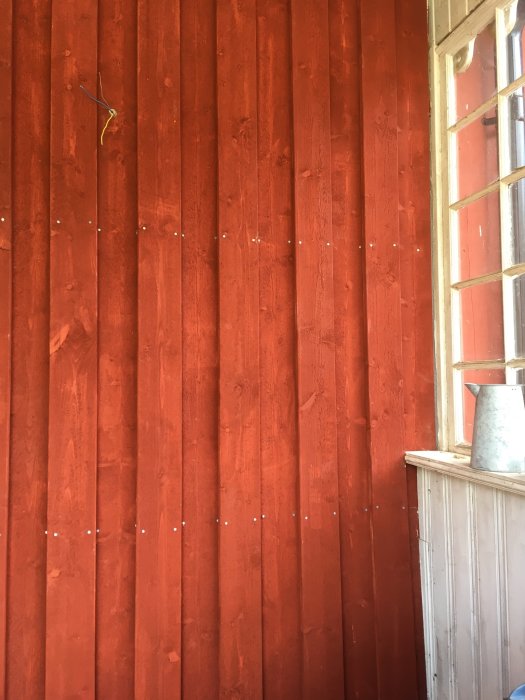 Nybyggd vägg med rödmålad träpanel och synlig del av ett fönster samt elledning.