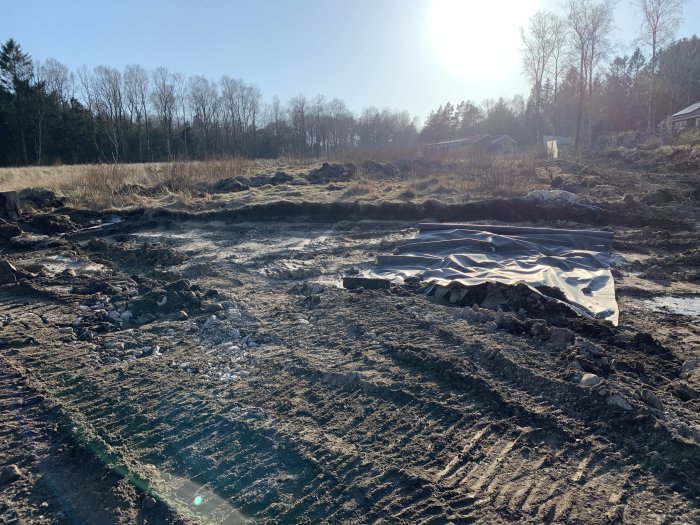 Uppgrävd byggplats med material och dränering i botten, solljus och skog i bakgrunden.