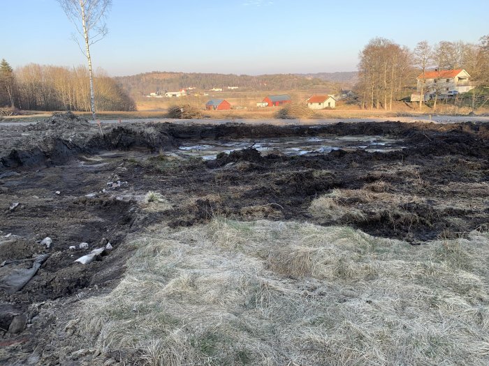 Grävmaskinen har påbörjat arbetet med att gräva ut för ett hus på en tom byggplats med frodig landsbygd i bakgrunden.