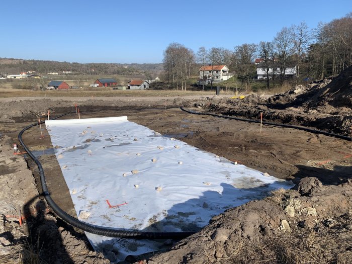 Utspridd vit duk och svart dräneringsrör på en grävd tomt med markerade gränser, i bakgrunden hus och natur.