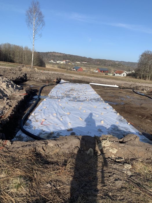 Uppgrävd byggplats för hus med nydragen dränering och vit markduk, omgivet av natur.