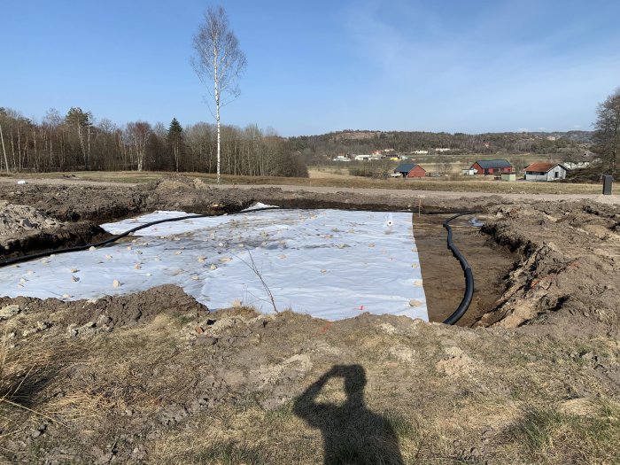 Utgrävningsarbete på en byggplats med synlig dräneringsslang, plastskydd på marken och omkringliggande landskap.