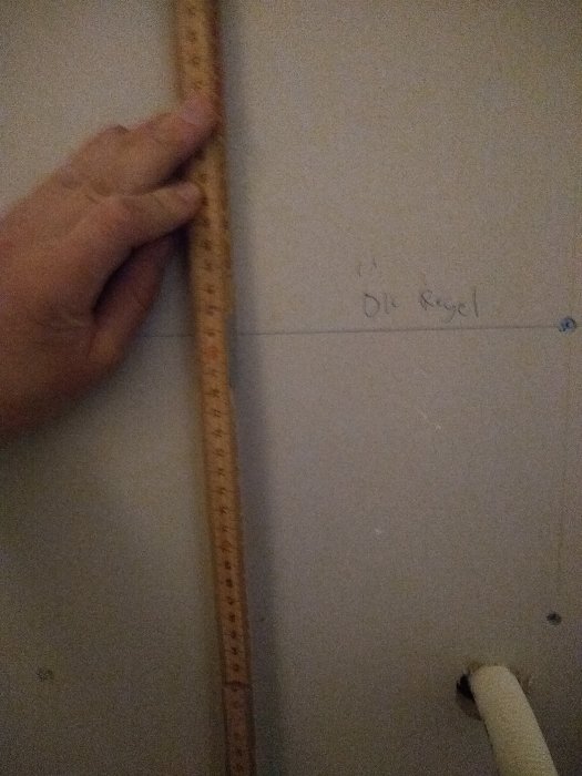 Hand håller en måttstock mot en vägg med markerad linje för framtida regel vid renovering.