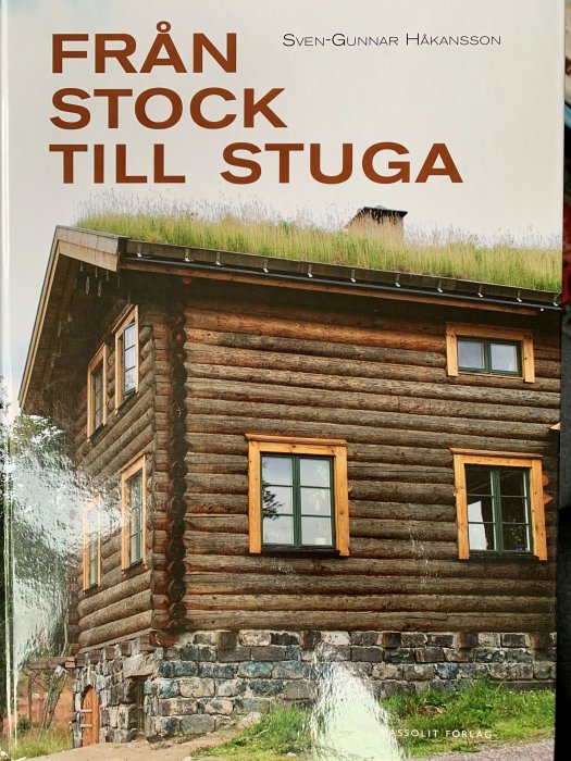 Omslaget på boken "Från Stock Till Stuga" visar en del av en timrad stuga med gräs på taket.