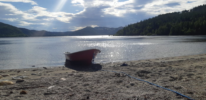 Röd roddbåt fastbunden vid strandkanten med glittrande sjö och skogklädda berg i bakgrunden.