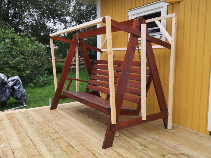 Hammock med trästomme och nybyggda reglar för att stödja ett plasttak, placerad på en trädäck framför ett gult hus.