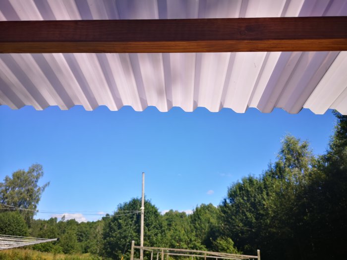 Vit plasttak med vågprofil fäst vid bruna träreglar ovanför hammock, med klarblå himmel och gröna träd i bakgrunden.