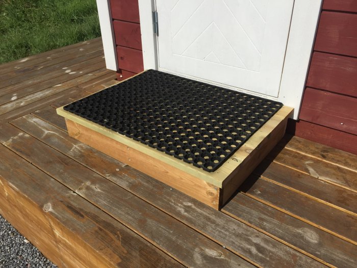 Ett nybyggt trästeg med svart dörrmatta framför en bastus dörr på en brygga vid stranden.