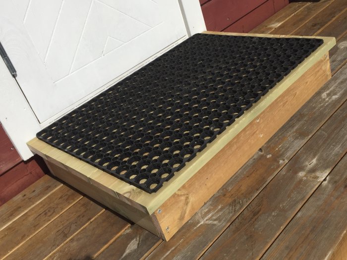 Ett nyinstallerat trästeg med gummimatta framför en dörr, placerat på en träbrygga i solljus.