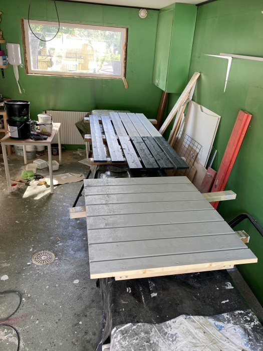 Nybyggd vindslucka av trä rankad på ett bord i ett rörigt garage med gröna väggar.
