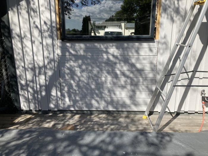 Nyinstallerad vit panel under fönster på garage med stege till höger och trädskuggor på marken.