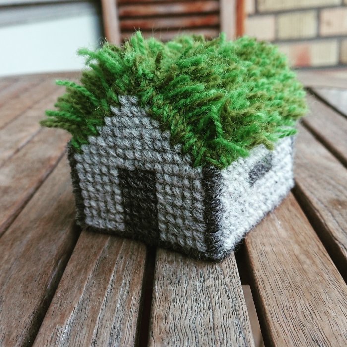 Handstickad miniatyr av ett hus med grönt tak som ligger på ett träbord utomhus.