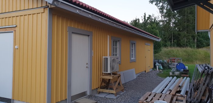 Gul panelbyggnad med röd takavvattning, luftvärmepump och byggmaterial i förgrunden.