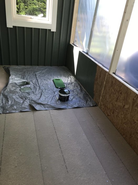 En halvfärdig renovering med oskyddade väggar, öppna fönsterramar och golvspån som delvis är lagt på ett rum under konstruktion.