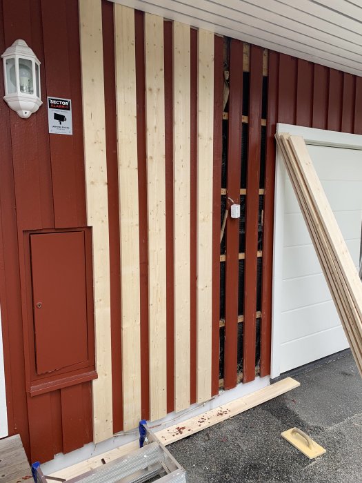 Renoveringsarbete på en husvägg med ny panel, ofärdig med synliga stolpar och verktyg på marken.