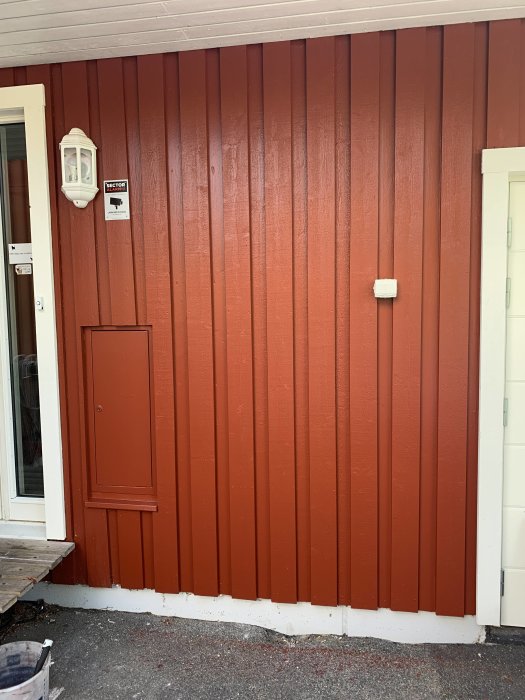 Rödmålad yttervägg med ny panel och vita lister där en inbyggd skåpdörr syns, efter renovering.