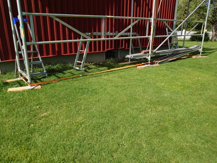 Ställning uppställd mot röd byggnad med förstärkta stödben på träplankor på gräs.