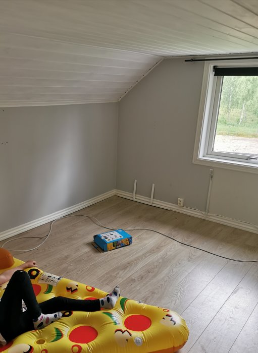 Renoverat barnrum med lutande tak, grå väggar, laminatgolv och ett fönster.