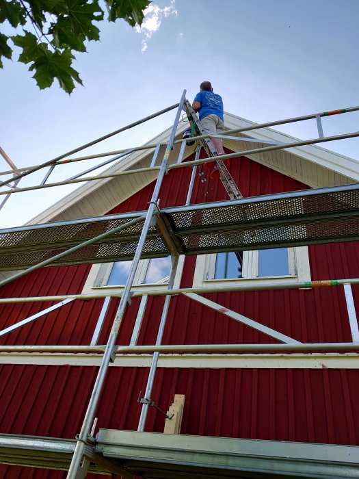 Person som arbetar på stege stödd mot byggnadsställning vid rött hus.