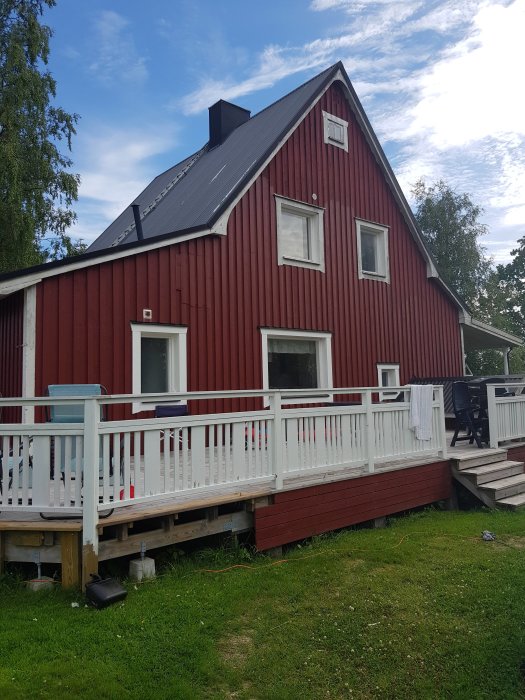 Rött trähus med svart tak och veranda omgiven av grönt gräs och träd mot blå himmel.