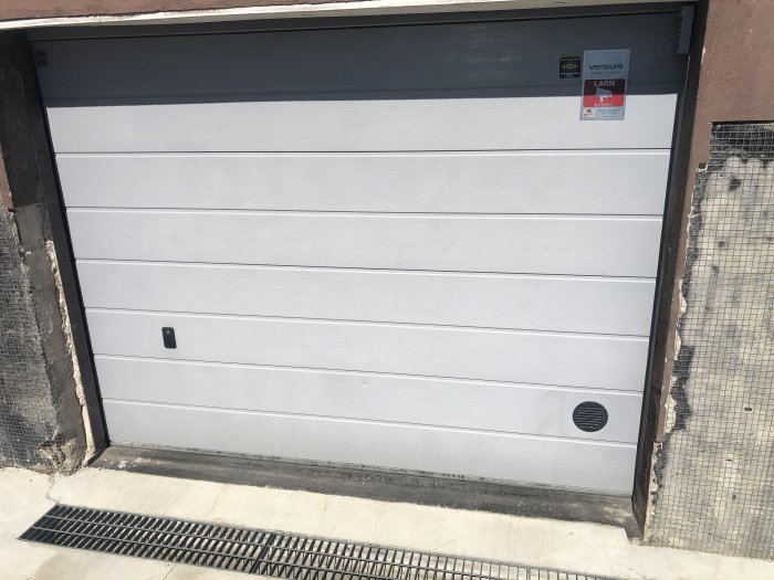 Stängd garageport med nyligen installerad tilluftsventil och larmdekal, i betongvägg och trottoar framför.