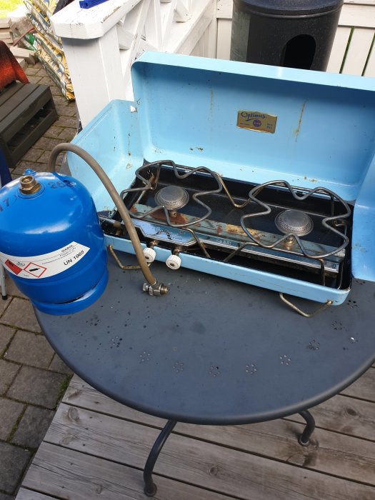 Nytt blått gasolkök för utomhusbruk anslutet till en äldre blå gasolflaska på ett grått bord.