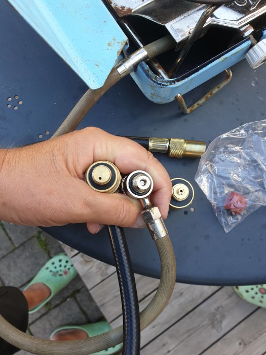 Hand som håller flera gasolflaskanslutningar bredvid ett blått gammalt gasolkök, på en träplattform utomhus.