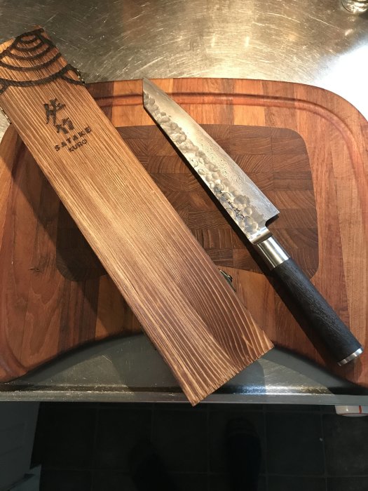 Japanskt kökskniv i damaskusstål bredvid sitt träfodral på en skärbräda.