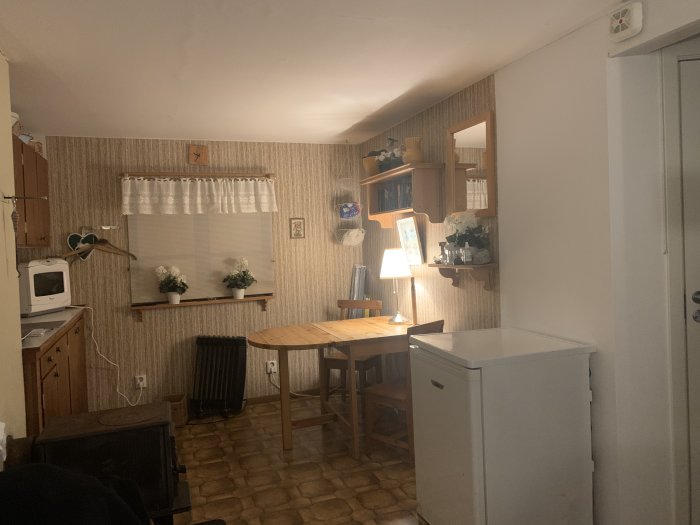 Interiör i lillstuga före renovering med teak köksskåp och vävtapet.