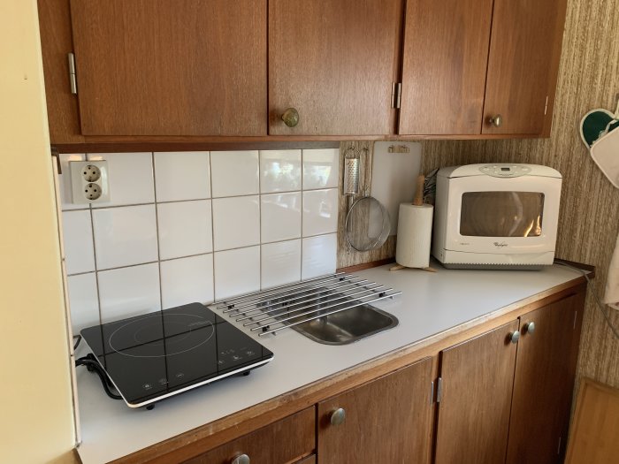 Äldre köksinteriör med teakskåp, vit kakelvägg, bärbar häll och mikrovågsugn, före renovering.