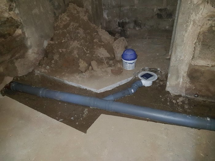 Grå avloppsrör delvis täckta med jord och cement i ett ofärdigt betongutrymme, med en hink Puts- och murbruk C.