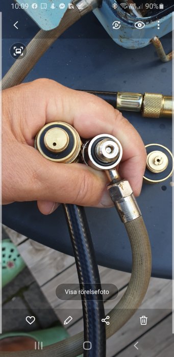 Hand som håller grillmunstycken för jämförelse; ett svart (nytt) och ett grått (gammalt) mot en gasoltub.