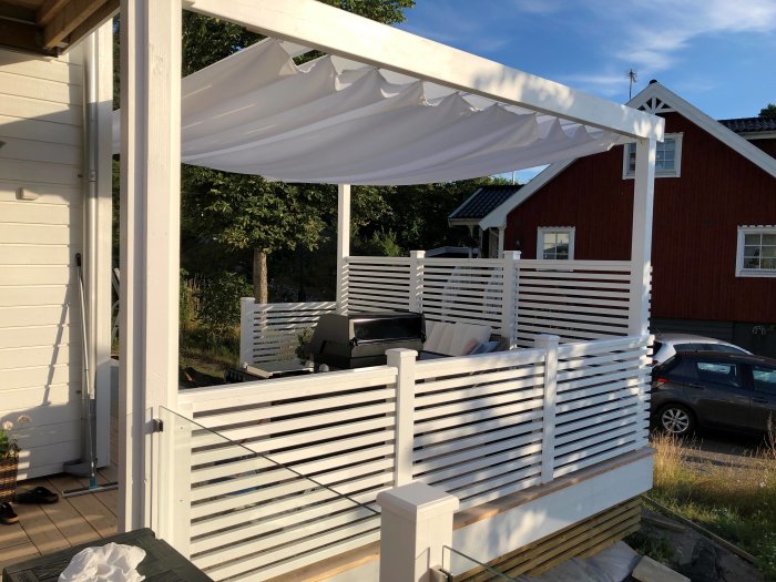 Vit pergola inredd med soltak på en träaltan, nära slutförda sittsteg med armågshyllor framför ett rött hus.