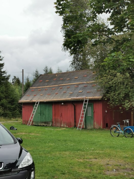 Ekonomibyggnad med rött trä och plåttak, borttagen plåt på ena sidan, stegar lutade mot väggen.