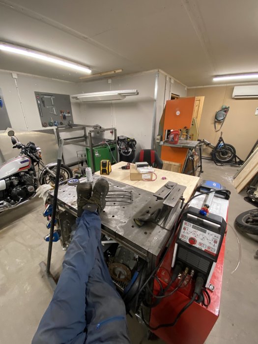 Vy över ett garage med sammanfogade arbetsbänkar i mitten, svetsutrustning, och verktyg, med en motorcykel i bakgrunden.