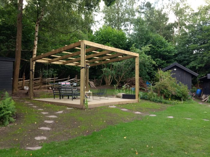 Trädgård med nyligen byggd obehandlad träpergola, stenstig framför och grönska i bakgrunden.