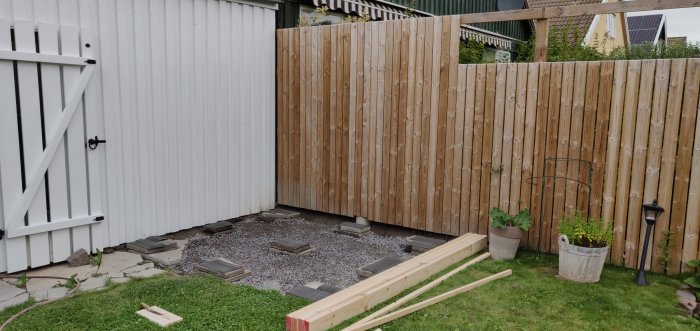 Hörn av en trädgård med vit carportdörr till vänster, träplank bakom gruslagd yta med stenplattor och träreglar på marken.