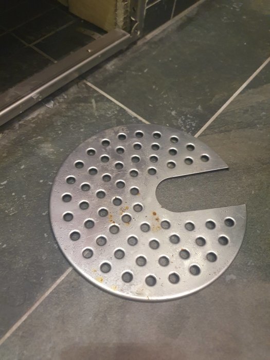 Rostangripen golvbrunnssil i metall med små rostfläckar placerad på klinkergolv.