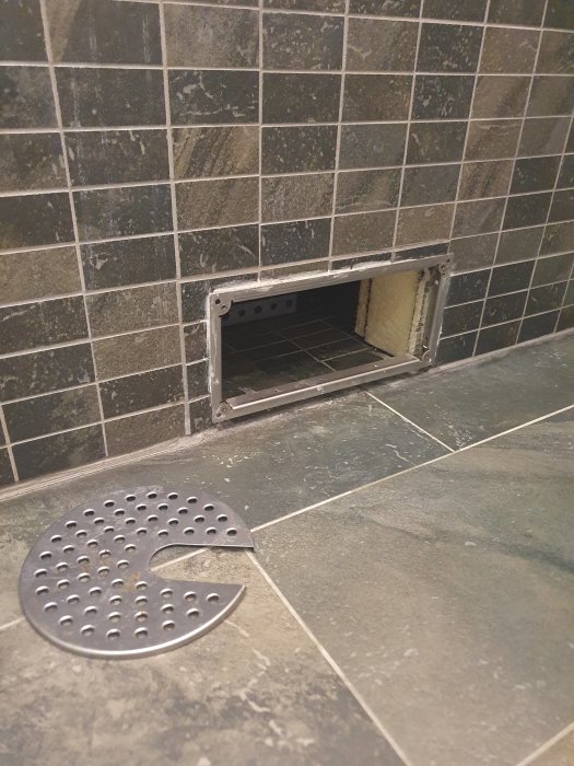 Rostangripen golvbrunn under inbyggt badkar, inspektionslucka öppen, och sil bredvid på klinkergolv.