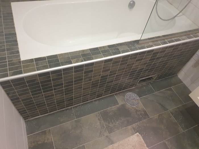 Badrum med grå klinkers, inbyggt badkar och rostangripen golvbrunn synlig genom inspektionslucka.