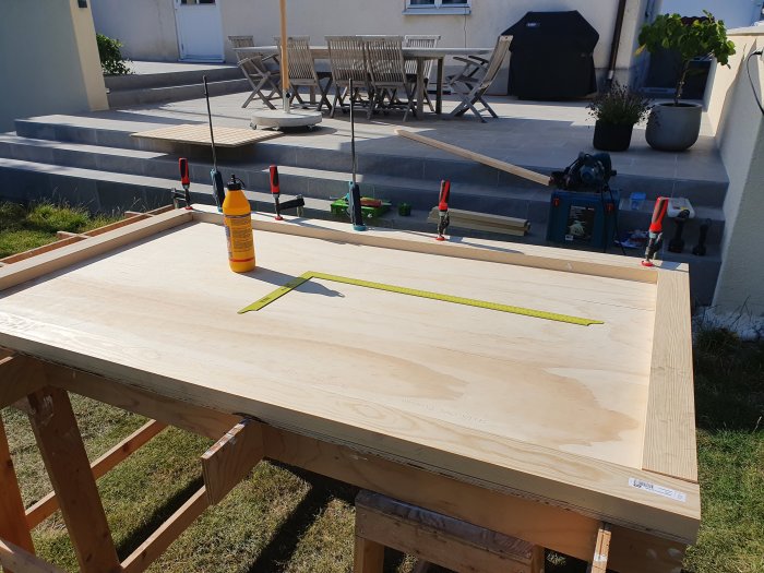 En dörr under konstruktion på ett arbetsbord utomhus med spårad plywood och verktyg, omgiven av en trädgård och uteplats.