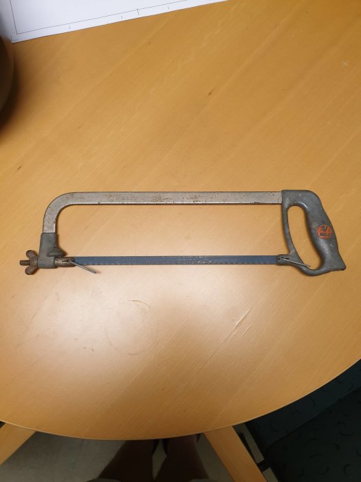 En gammal bågfil av metall med bladet monterat, ligger på ett ljust träbord, eventuellt köpt begagnad.